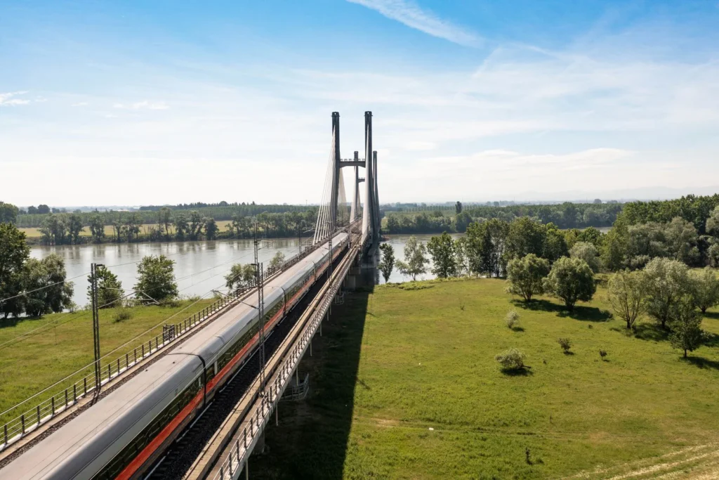 Train Frecciarossa traversant un pont au-dessus d’un fleuve entouré de verdure sous un ciel bleu.