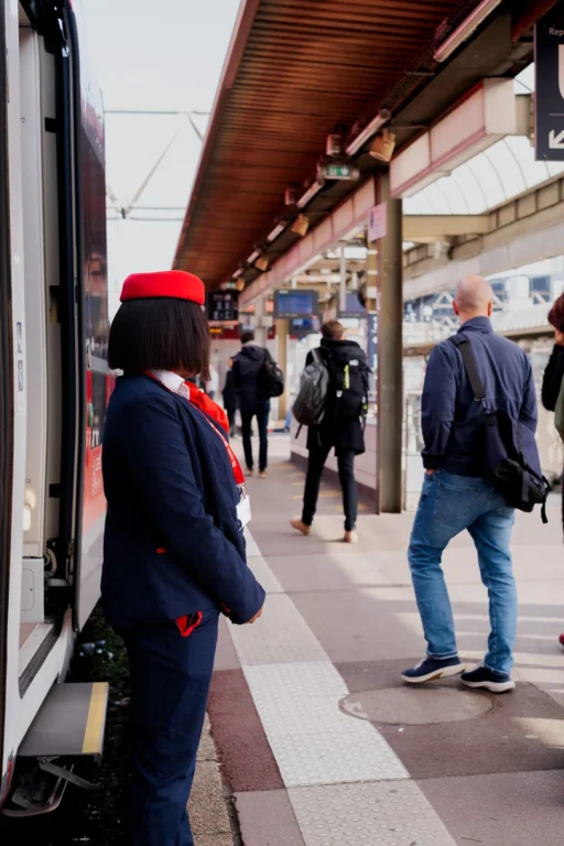 Agent de train en uniforme bleu et chapeau rouge surveillant l’embarquement des passagers sur le quai d’une gare.