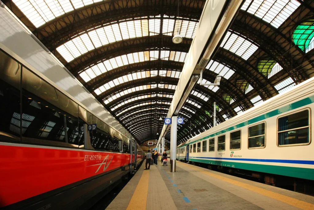 Vue intérieure de la gare de Milan avec un train Eurostar rouge et argent à gauche et un train régional Trenitalia à droite sous la grande verrière métallique