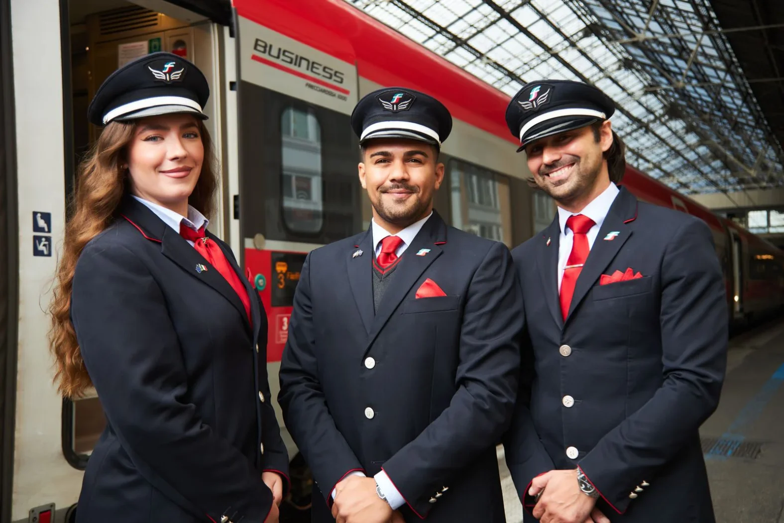 Trois agents de bord Trenitalia en uniforme bleu marine et cravate rouge posant souriants devant un train rouge et argent dans une gare couverte.
