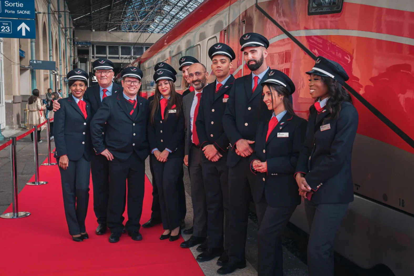Équipe de personnel Trenitalia en uniforme bleu marine et cravate rouge posant ensemble sur un tapis rouge devant un train Frecciarossa dans une gare couverte.