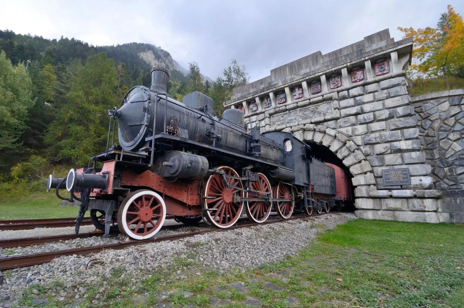 Locomotive à vapeur noire et rouge sortant d’un tunnel en pierre, entourée de verdure et de montagnes en arrière-plan, sous un ciel nuageux.