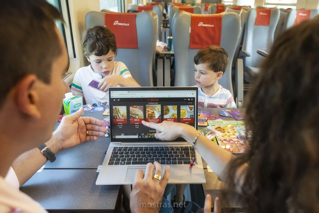 Famille à bord d’un train Trenitalia, les parents choisissent un menu EasyBistrò sur un ordinateur portable pendant que les enfants jouent à table