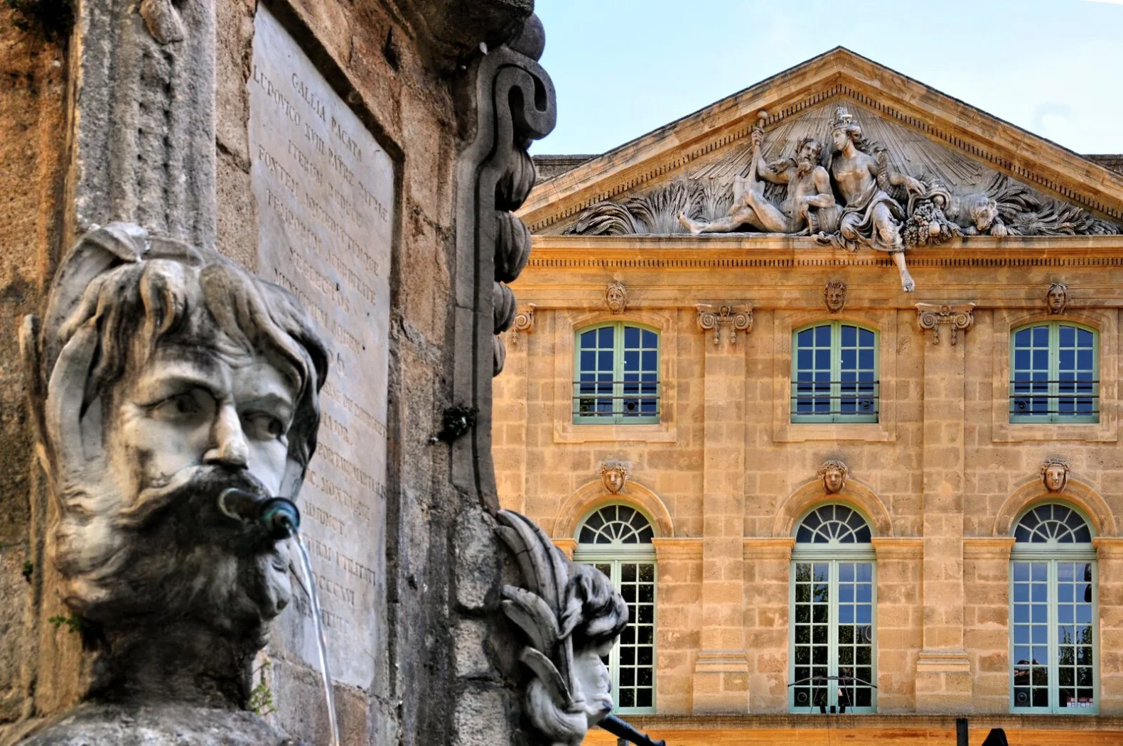Détail d’une fontaine sculptée à Aix-en-Provence avec en arrière-plan la façade d’un bâtiment classique orné de statues et bas-reliefs.