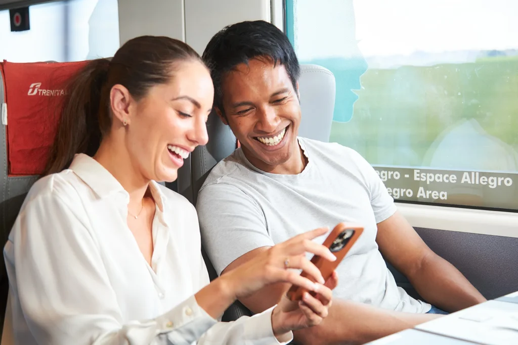 Deux passagers souriants regardant un téléphone ensemble dans un train Frecciarossa, assis côte à côte près de la fenêtre.