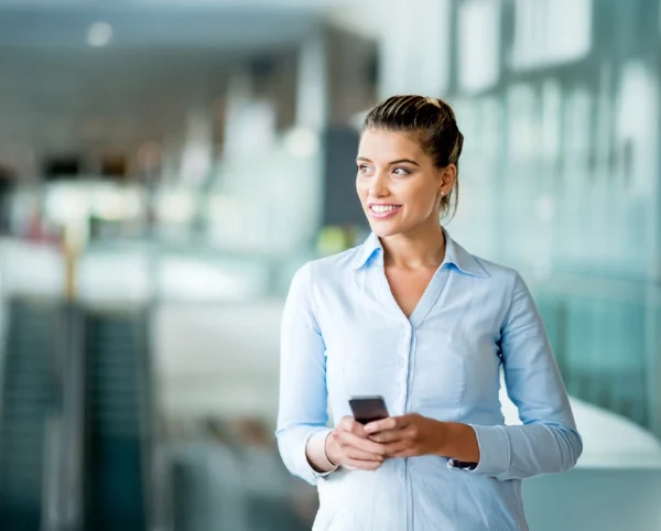 Femme souriante en chemise claire tenant un smartphone dans un espace moderne et lumineux