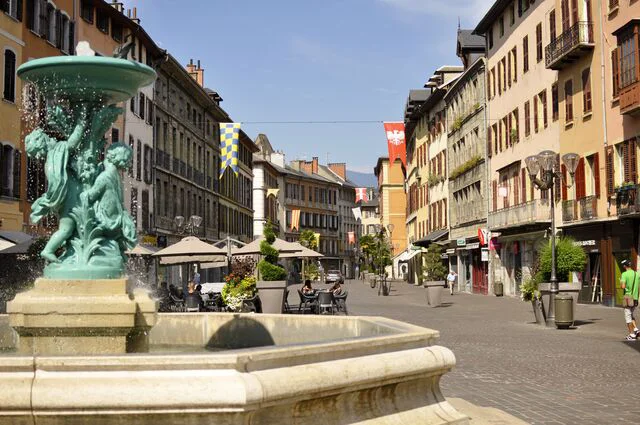Fontaine et terrasse de café dans la vieille ville d’Annecy, entourée de bâtiments colorés et de rues pavées.