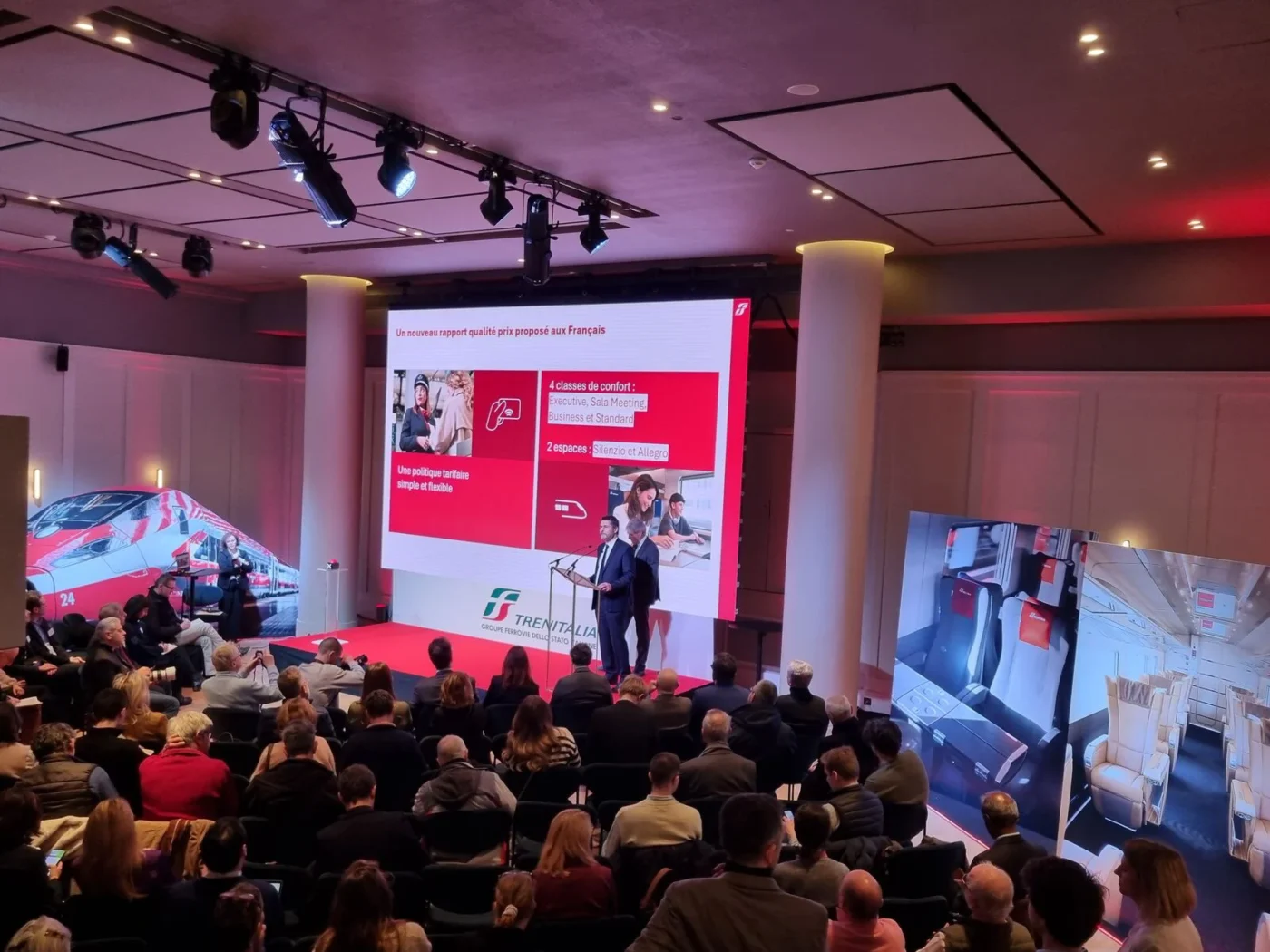 Conférence Trenitalia présentant les services Frecciarossa, avec un intervenant sur scène devant un grand écran rouge et blanc et un public attentif dans la salle.