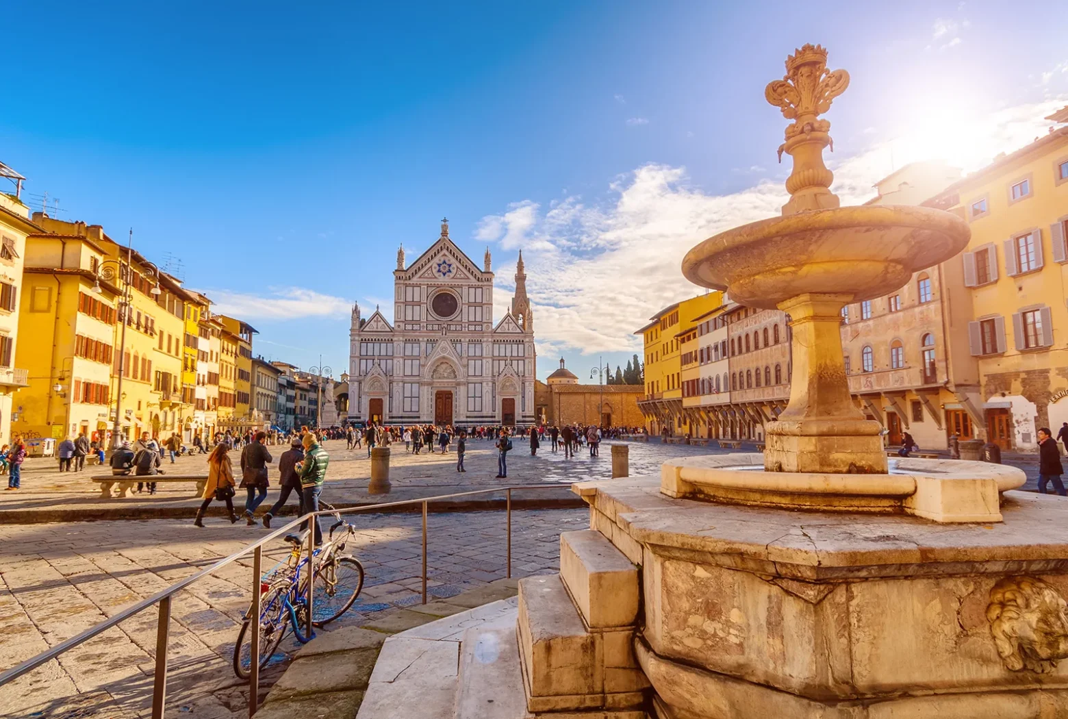 Vue ensoleillée de la Piazza Santa Croce à Florence, avec la basilique Santa Croce au fond, une fontaine en marbre au premier plan et des passants sur la place.