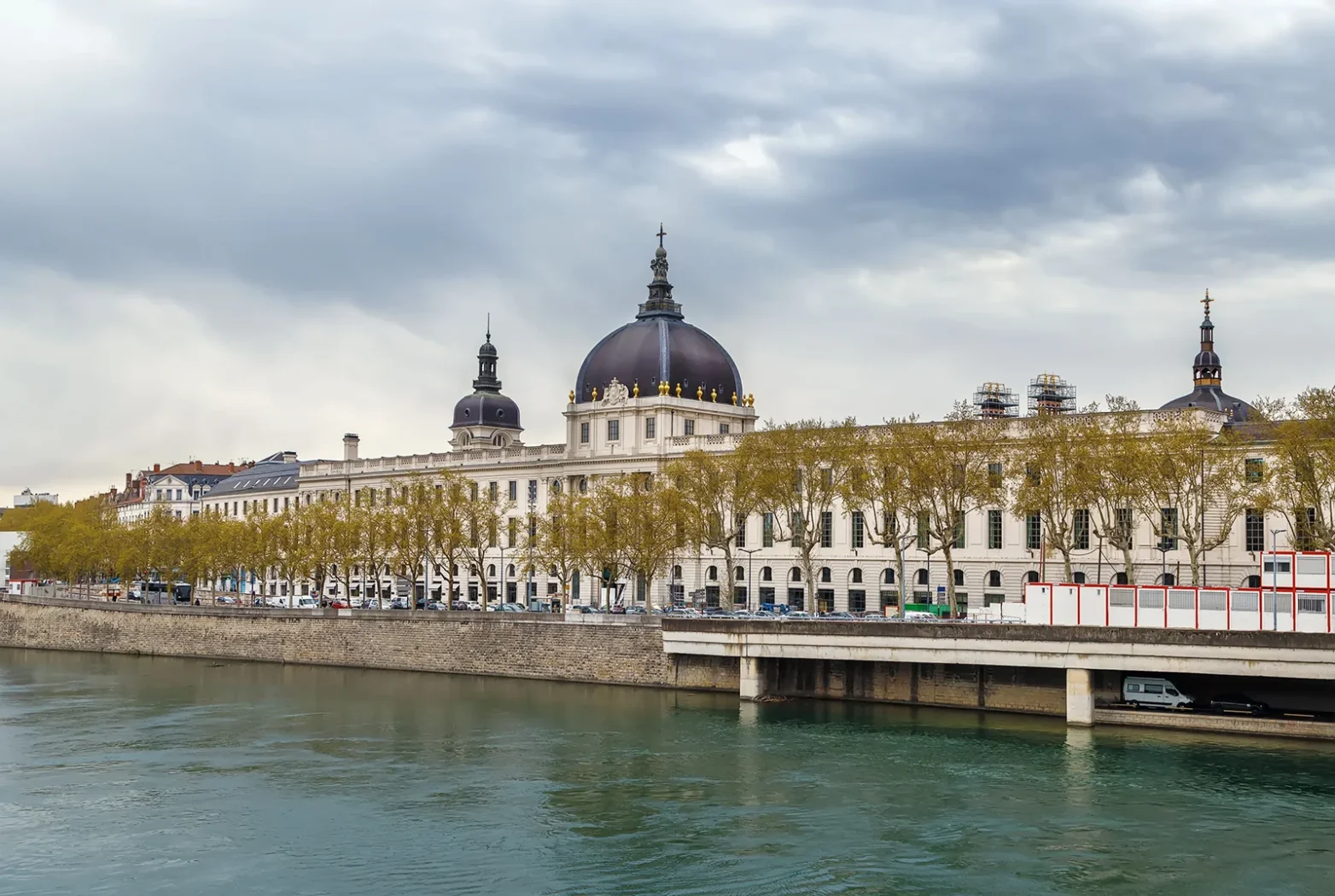 Vue du majestueux Hôtel-Dieu de Lyon au bord du Rhône, avec son dôme emblématique et les arbres alignés le long du quai.