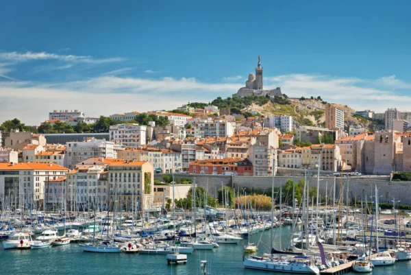 Vue panoramique du Vieux-Port de Marseille avec la basilique Notre-Dame de la Garde dominant la ville sous un ciel bleu.