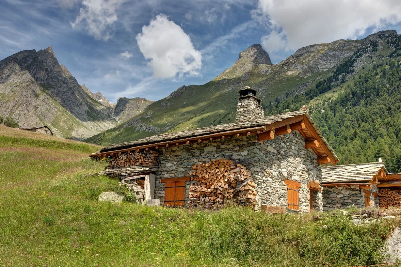 Chalet en pierre typique du Val d’Aoste avec bois empilé et montagnes verdoyantes en arrière-plan sous un ciel bleu.