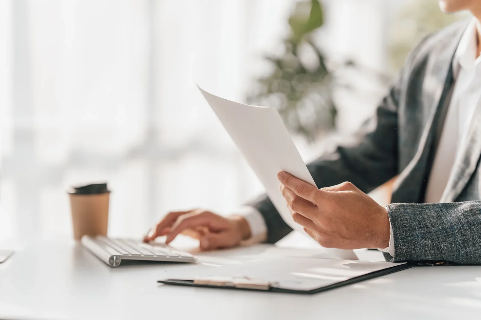Personne en costume examinant un document papier tout en utilisant un clavier d’ordinateur sur un bureau lumineux, avec un gobelet de café en arrière-plan.