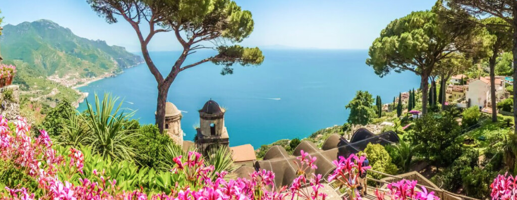 Vue panoramique sur la côte amalfitaine avec mer turquoise, collines verdoyantes, pins parasols et fleurs roses en premier plan.