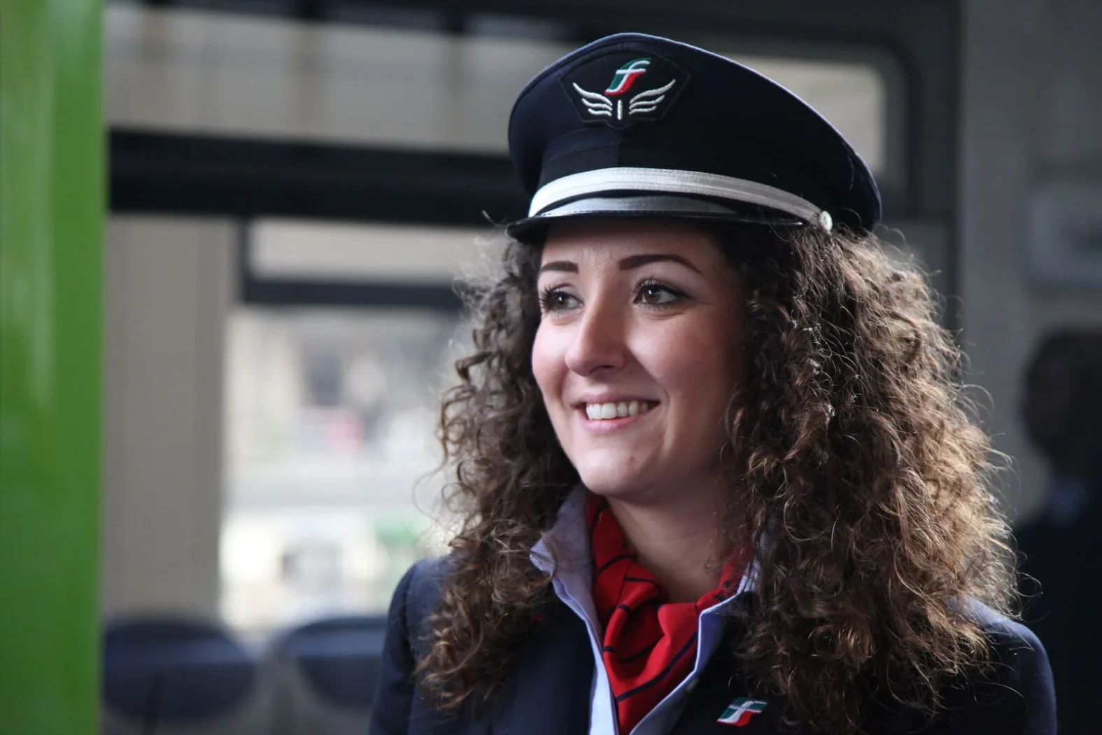 Personnel Trenitalia souriante portant un uniforme bleu marine, une cravate rouge et une casquette avec le logo de la compagnie.