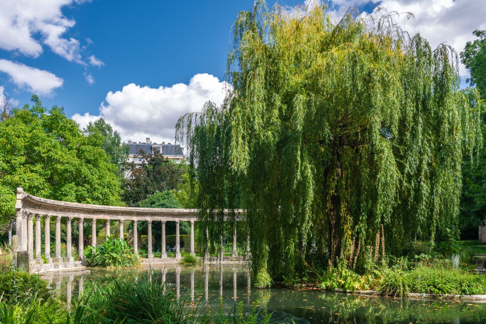Jardin verdoyant avec un étang, une colonnade en pierre et un grand saule pleureur.