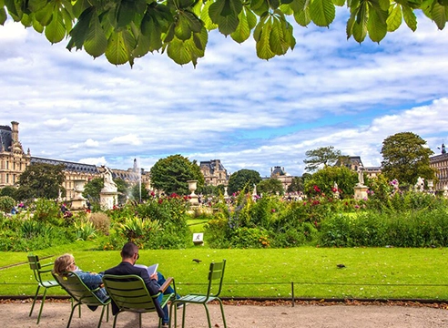 Couple assis sur des chaises vertes dans le jardin des Tuileries, avec vue sur le Louvre et les parterres fleuris.