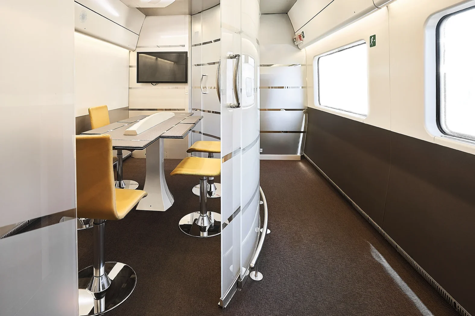 Salle de réunion moderne à l’intérieur d’un train, avec table, chaises jaunes et grand écran mural.