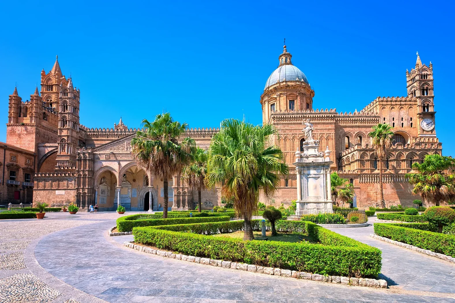 Façade majestueuse de la cathédrale de Palerme entourée de palmiers et de jardins, sous un ciel bleu éclatant.