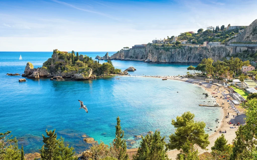 Vue aérienne de la plage et de l’île d’Isola Bella à Taormine, entourée d’une mer bleu azur et de falaises verdoyantes.