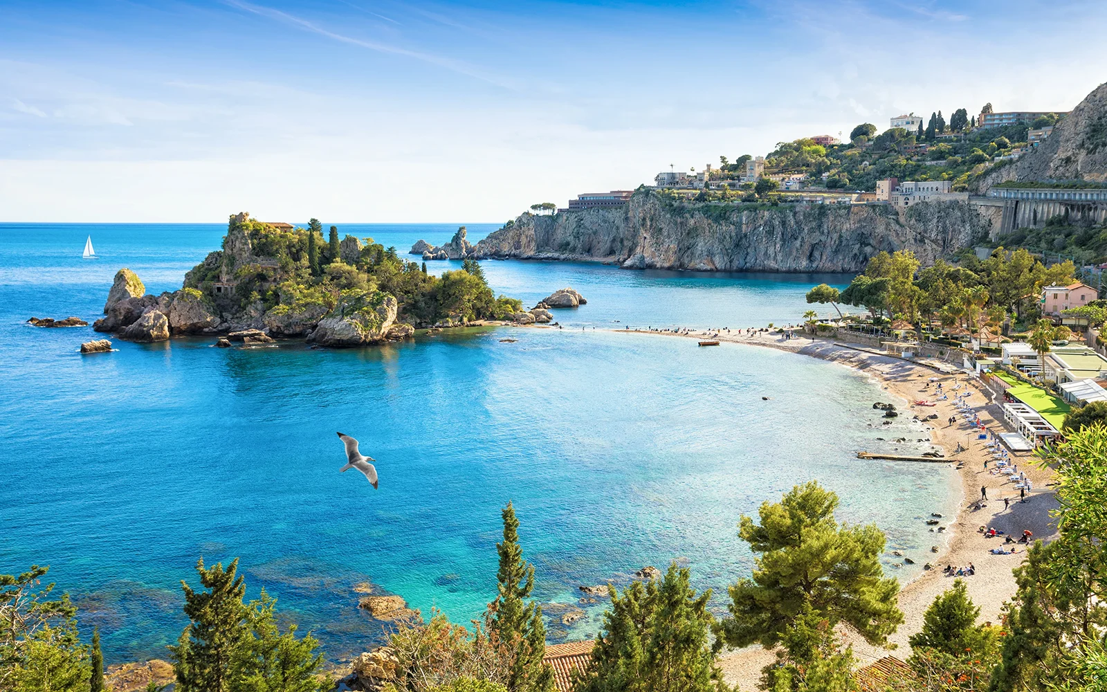 Vue aérienne de la plage et de l’île d’Isola Bella à Taormine, entourée d’une mer bleu azur et de falaises verdoyantes.