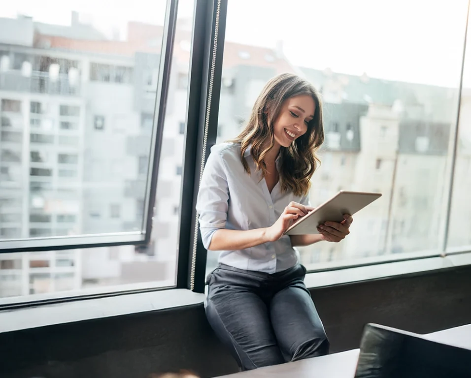 Femme souriante utilisant une tablette près d’une grande fenêtre dans un environnement professionnel lumineux