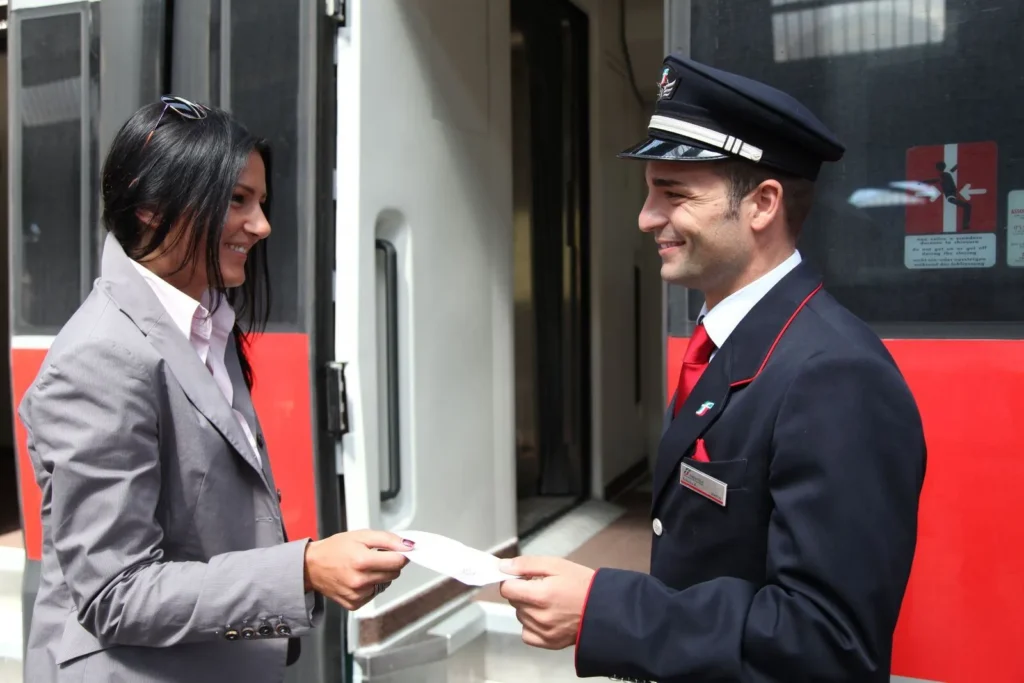 Un agent de train en uniforme souriant contrôle le billet d’une passagère sur le quai.