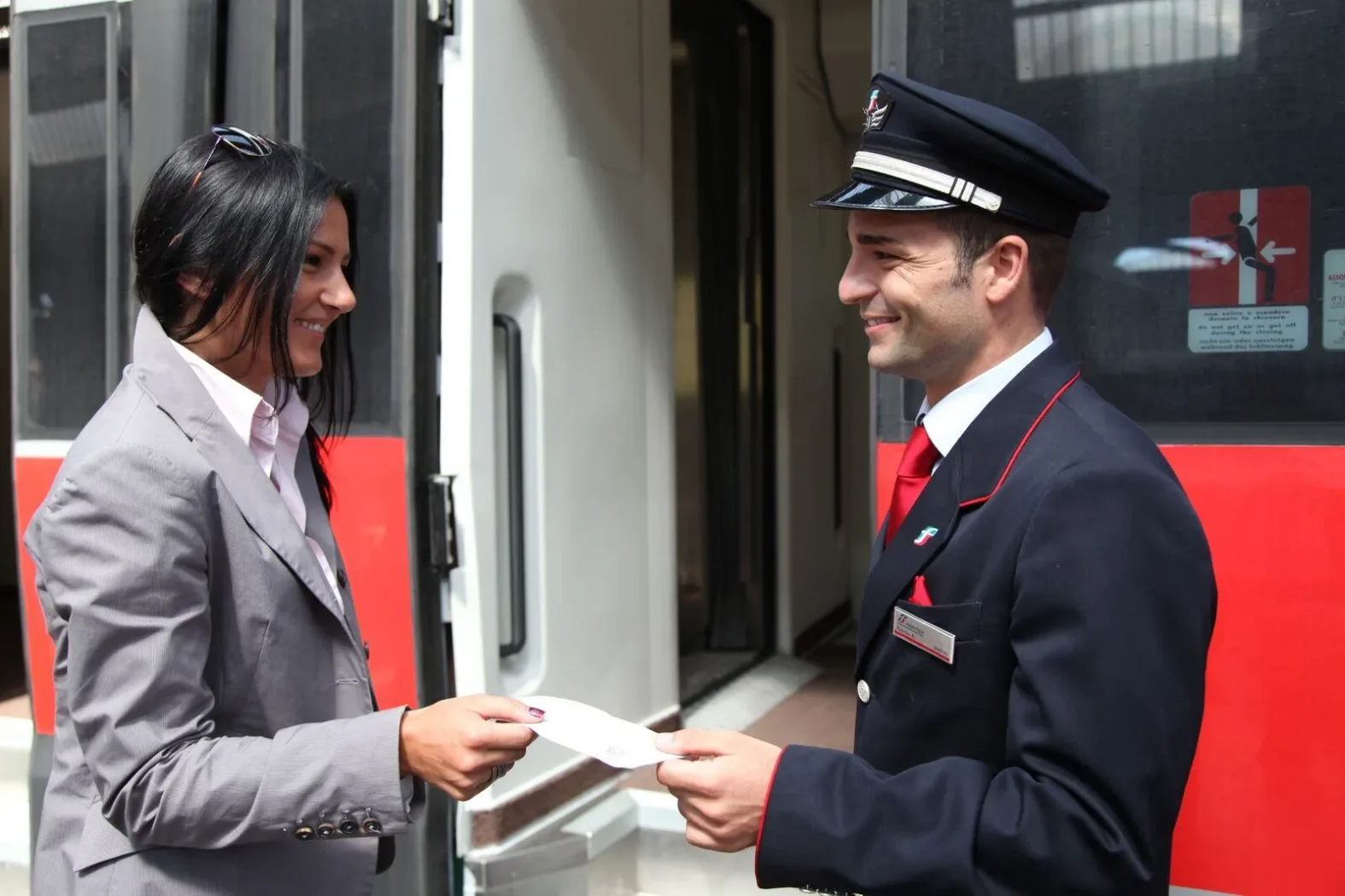 Un agent de train en uniforme souriant contrôle le billet d’une passagère sur le quai.