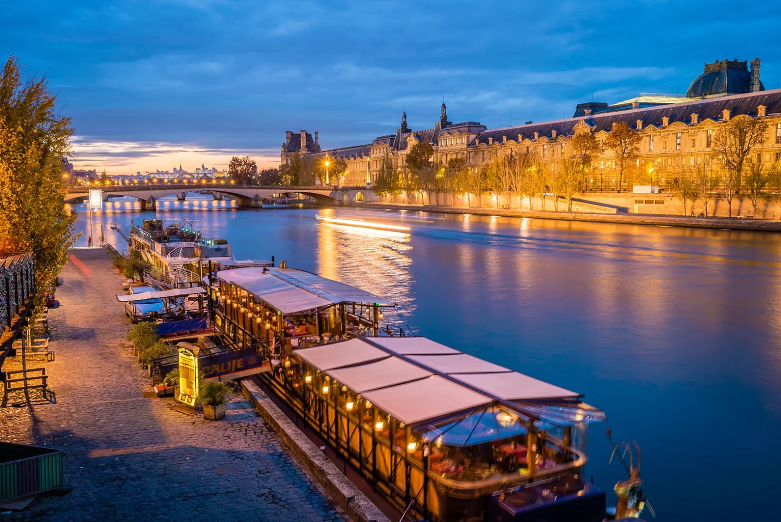 Vue nocturne des quais de Seine avec péniches illuminées et bâtiments historiques en arrière-plan.