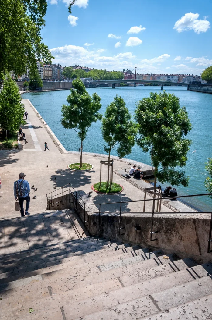 Vue ensoleillée des berges du Rhône à Lyon avec des passants, des arbres et un pont au loin sous un ciel bleu