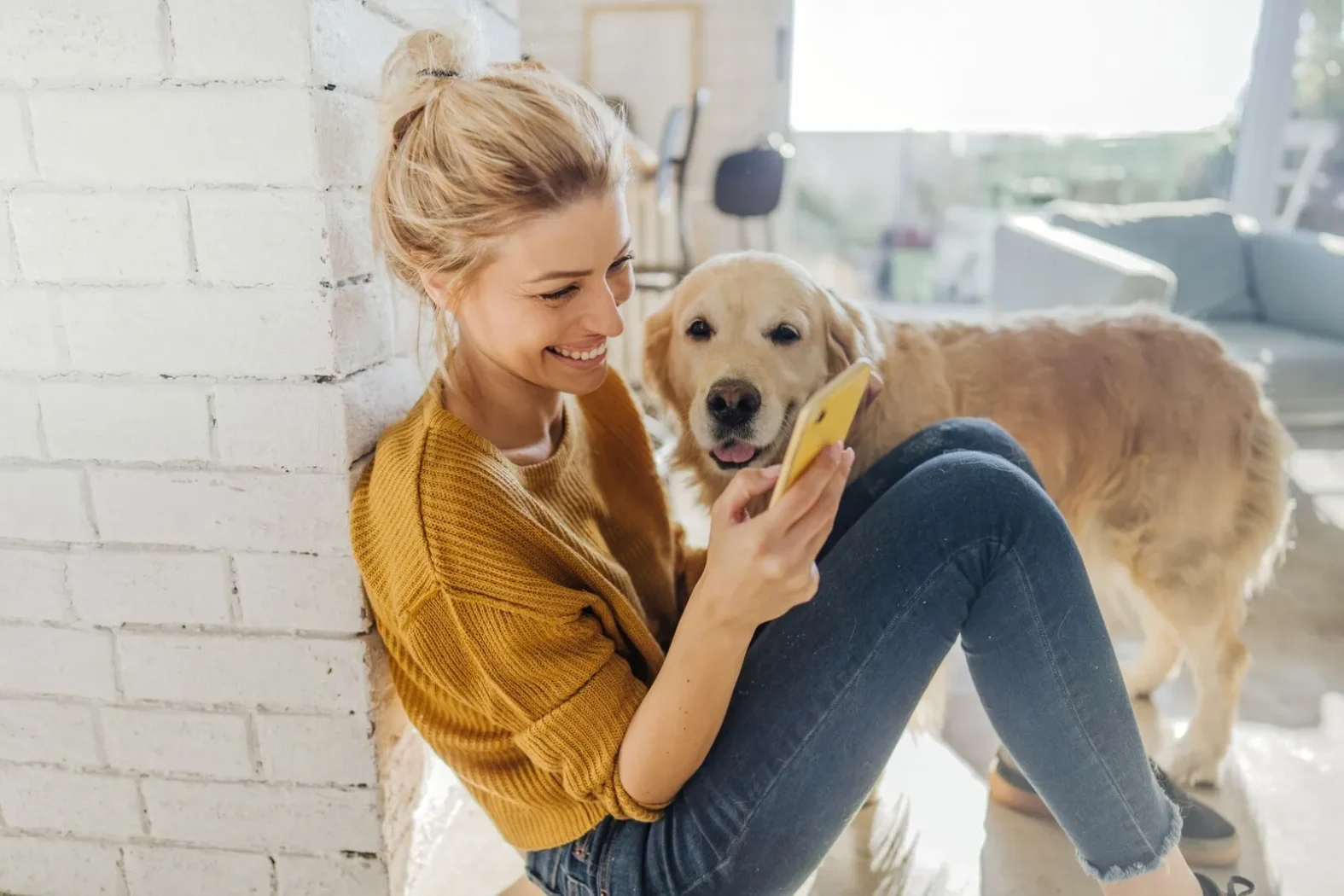 Femme souriante assise au sol avec son chien, regardant son téléphone portable dans un salon lumineux