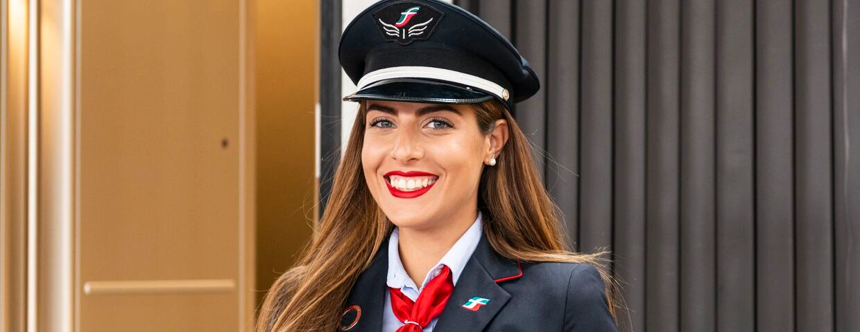 Contrôleuse Trenitalia souriante en uniforme bleu marine et cravate rouge tenant une tablette devant la porte d’un train.
