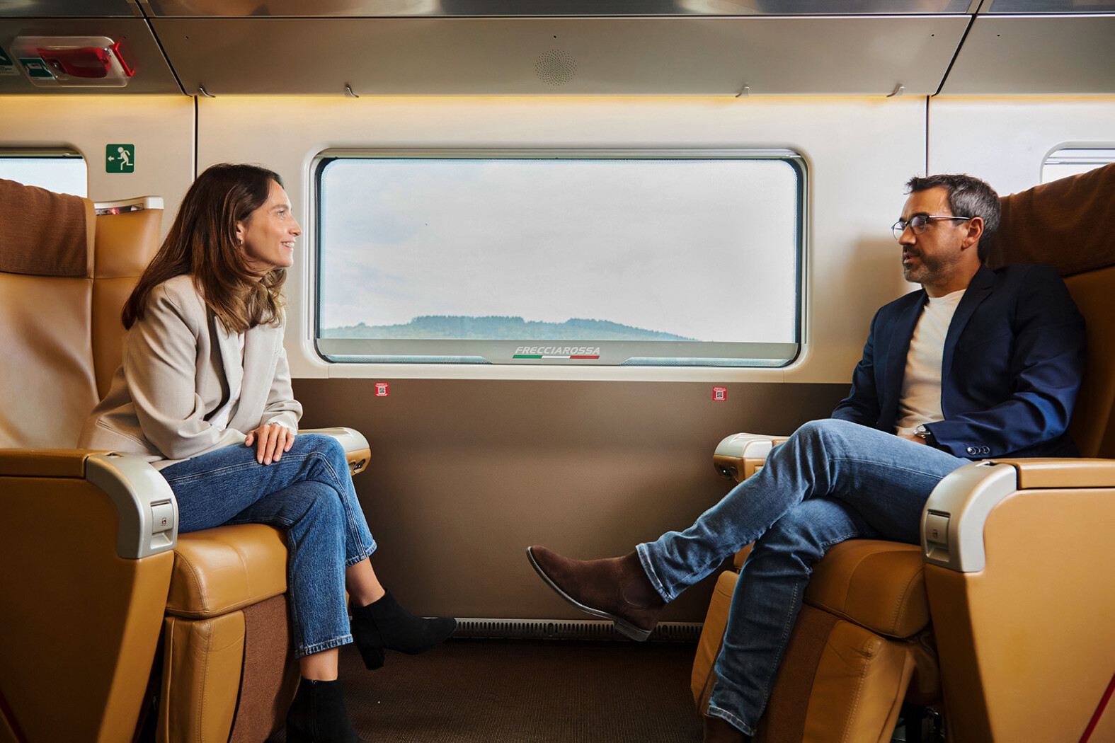 Deux passagers assis face à face dans un train Trenitalia Frecciarossa, discutant confortablement sur des sièges en cuir beige près d’une fenêtre.