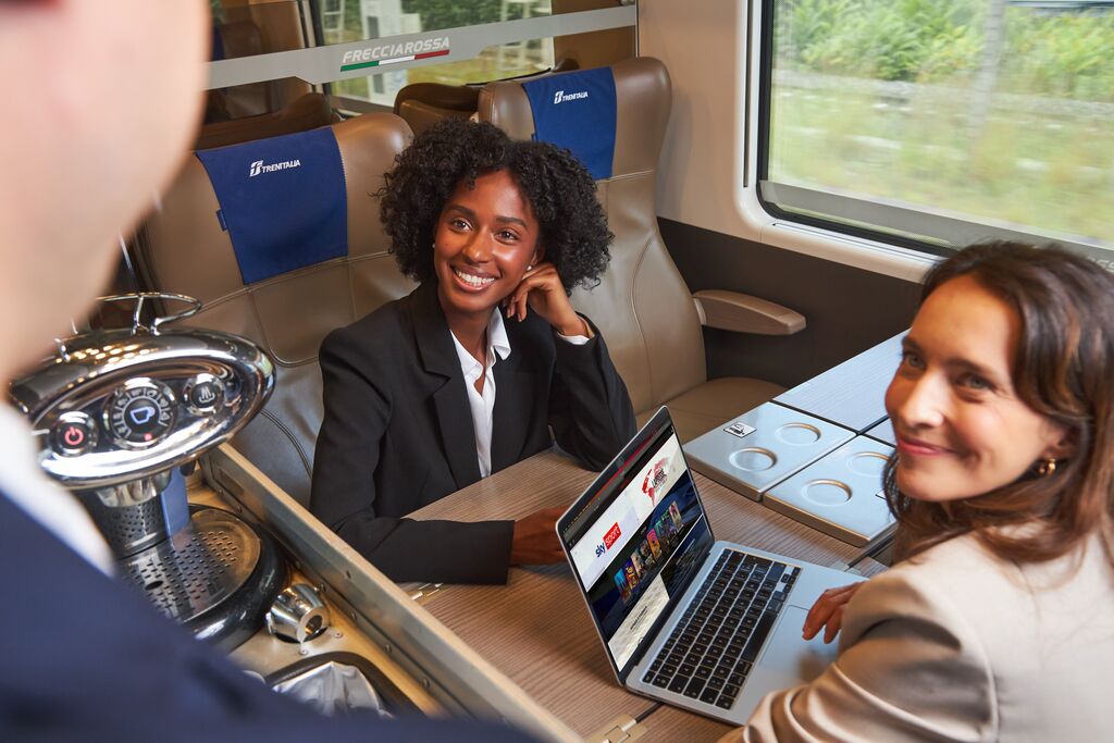 Deux femmes en tenue professionnelle discutant autour d’un ordinateur portable dans un compartiment de train moderne.