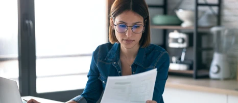 Femme portant des lunettes lisant un document devant son ordinateur dans un bureau lumineux.