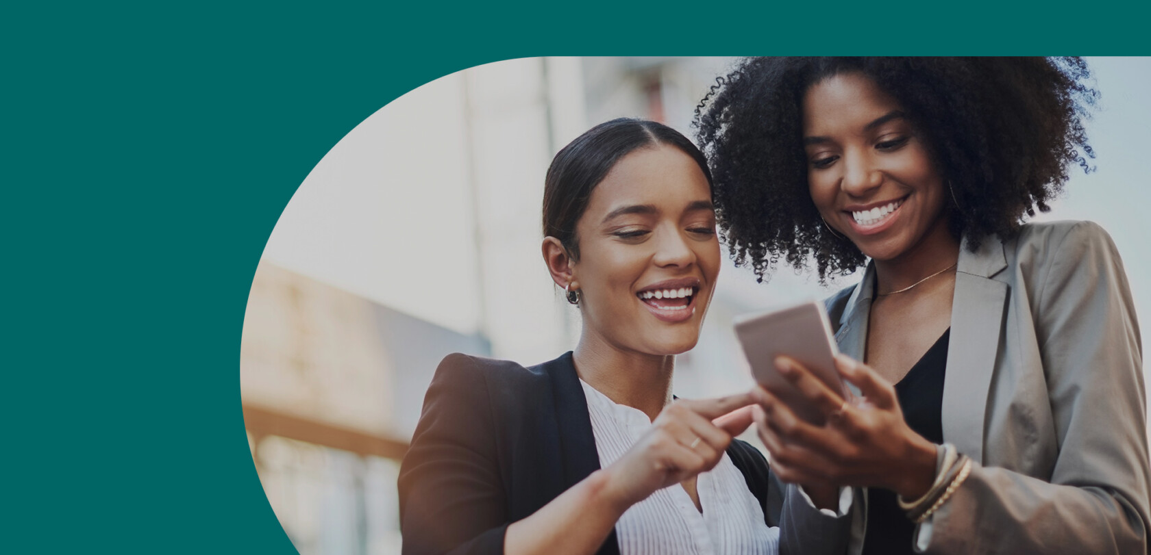 Deux femmes souriantes en tenue professionnelle regardent ensemble un téléphone portable.